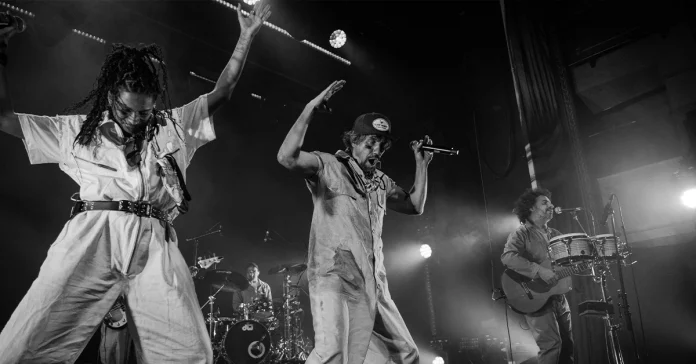 Artistas en el escenario bailando y cantando con monos de trabajo claros, acompañados por banda y luces de concierto