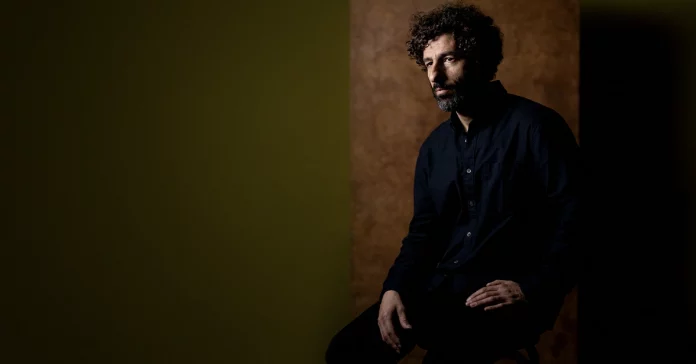 Hombre de barba y pelo rizado oscuro sentado en actitud relajada, mirada directa e intensa, camisa azul marino, fondo verde oscuro suave